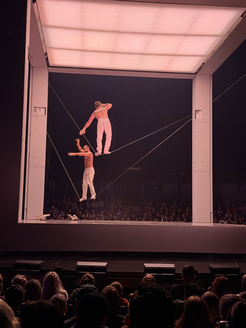 Two acrobats performing on a high wire suspended in a bright square frame above a dark arena