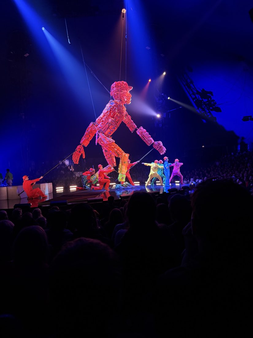 A giant mechanical puppet made of colorful interlocking pieces, suspended above acrobats in a darkened arena